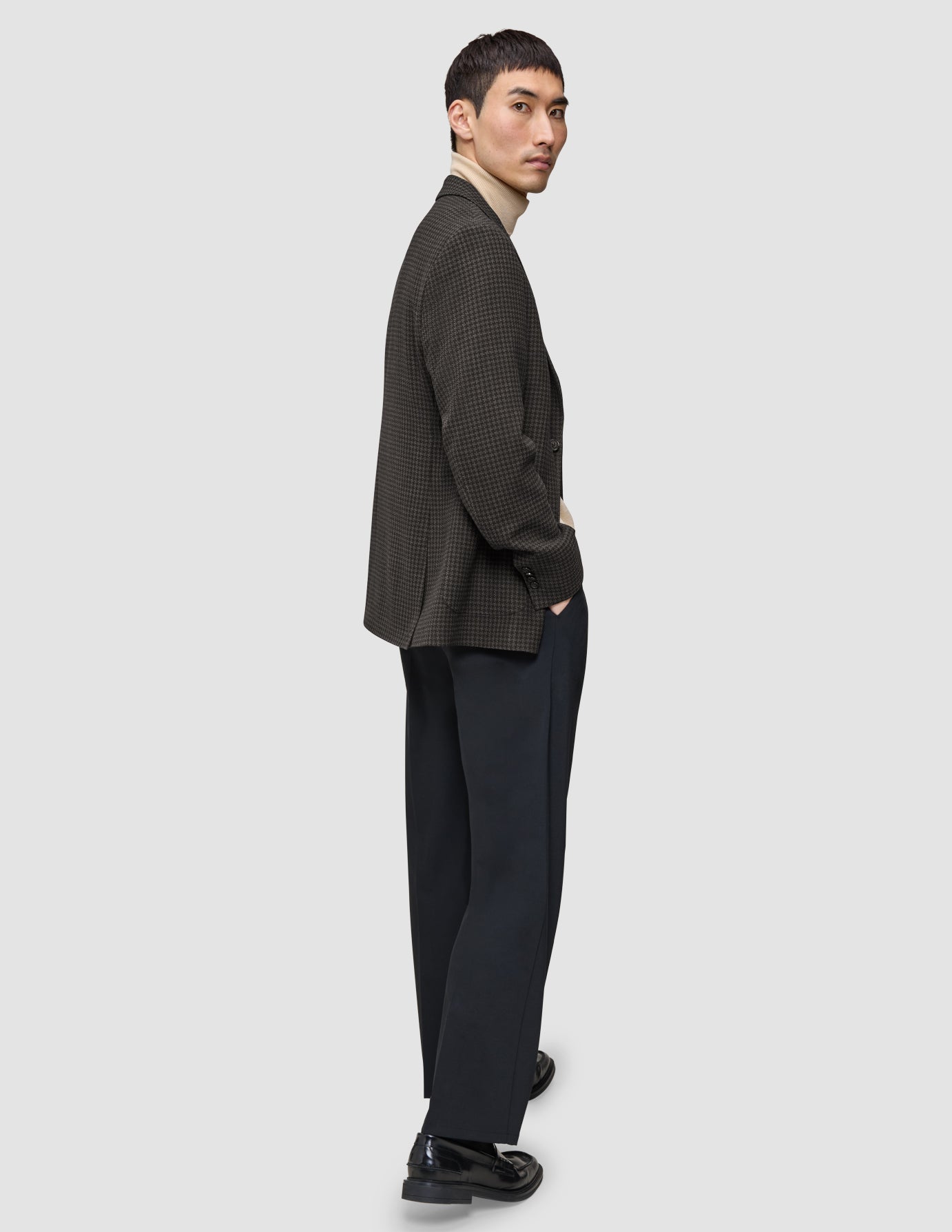 Ein Mann, der einen dunkelbraunen, karierten Timeless Wool Blazer Slim über einem beigen Rollkragenpullover und einer dunklen Hose trägt, blickt über seine Schulter.